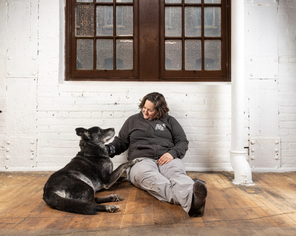 Pet photographer Robyn is sitting with her lab mix against a white brick wall under a brown paned window