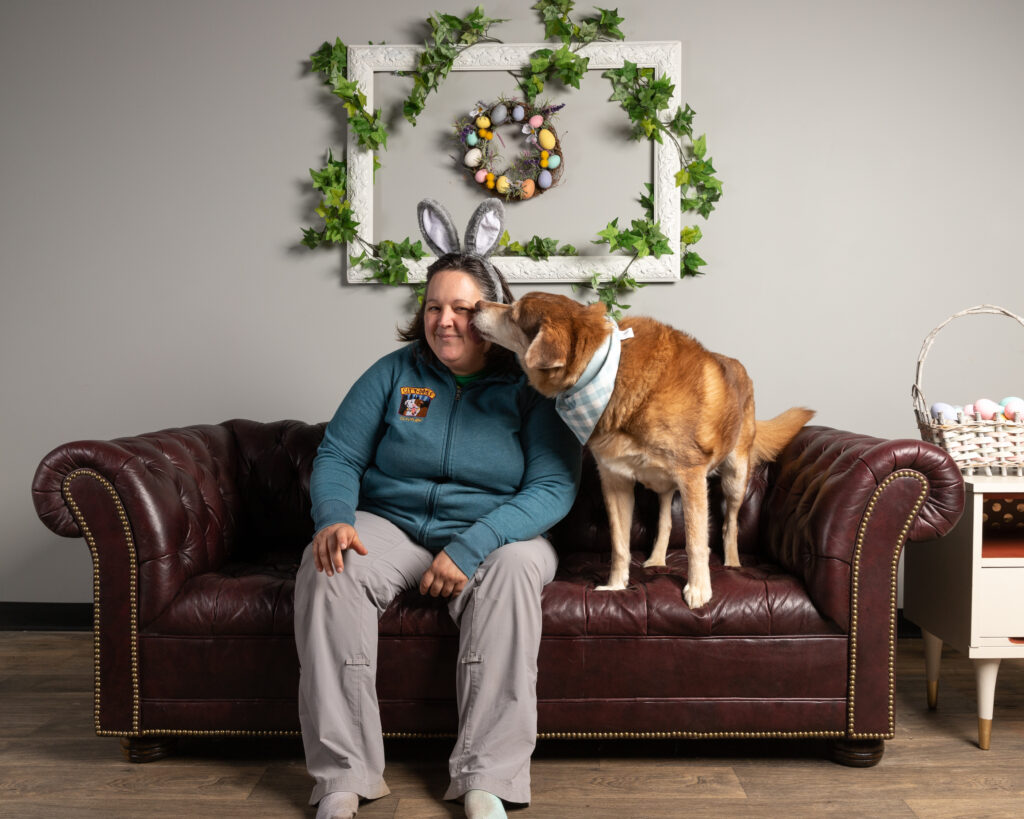 Pet photographer Robyn is sitting on a brown leather couch with a golden fluffy dog. On the wall is a white picture frame with ivy around it. Inside the frame is a wreath with Easter eggs attached. The dog is giving Robyn a kiss on the cheek.