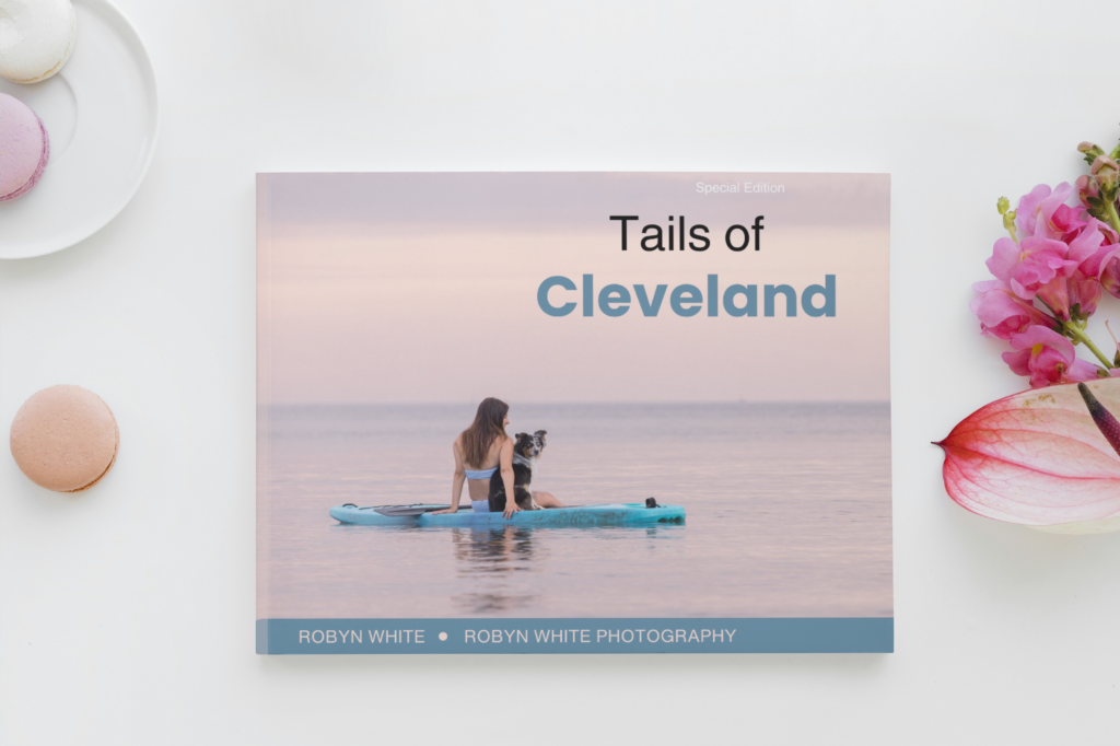 A mock up of a book titled Tails of Cleveland. There is a woman and a dog on a paddle board in Lake Erie.