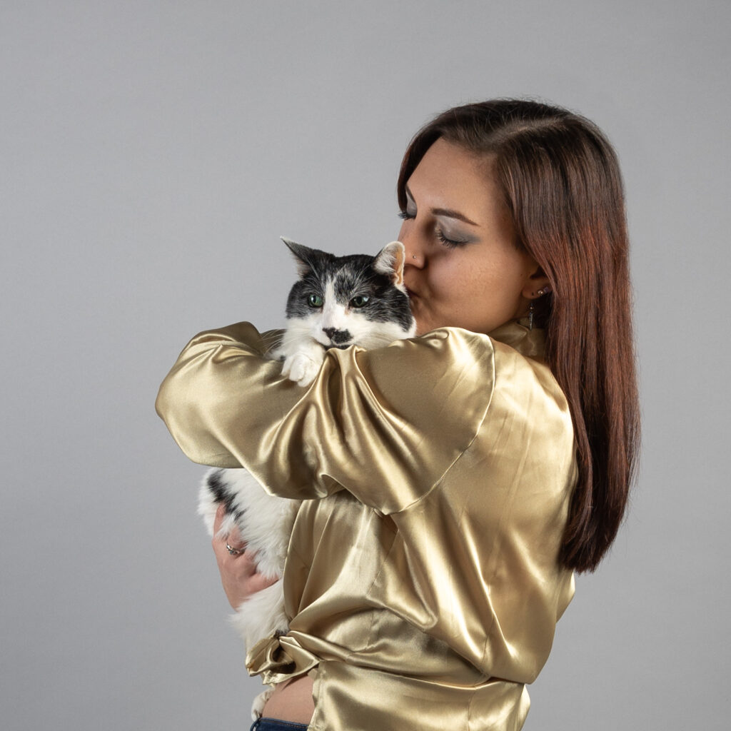 A woman wearing a gold shirt is holding and kissing her cat in front of a grey wall in a Cleveland Ohio studio
