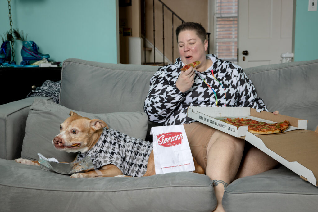 In-home photo session in Cleveland, Ohio with a senior pitbull and her mom lying on the couch. They are wearing matching houndstooth sweaters and eating burgers and pizza.