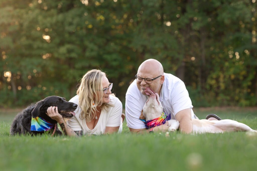 A man and woman are lying in the grass with their two dogs at the Cuyahoga National Park. One dog is kissing the man while the woman looks at them while stroking the chin of the other dog.